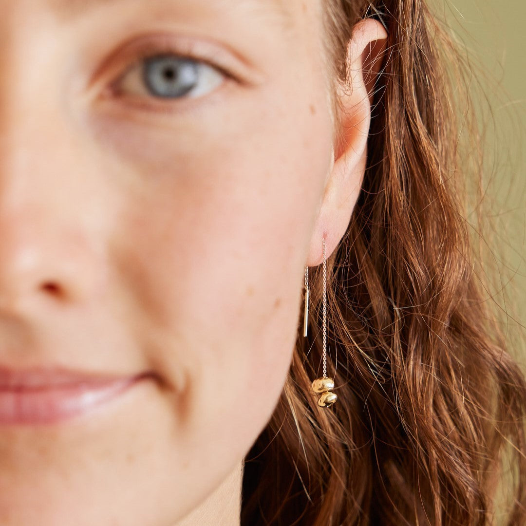 Halve gezicht van model met focus op de doorrijgoorbel die ze draagt: Oorhanger Essentials, 14 k gouden minimalistische steentjes aan een kettinkje welke je door je oren kan trekken.