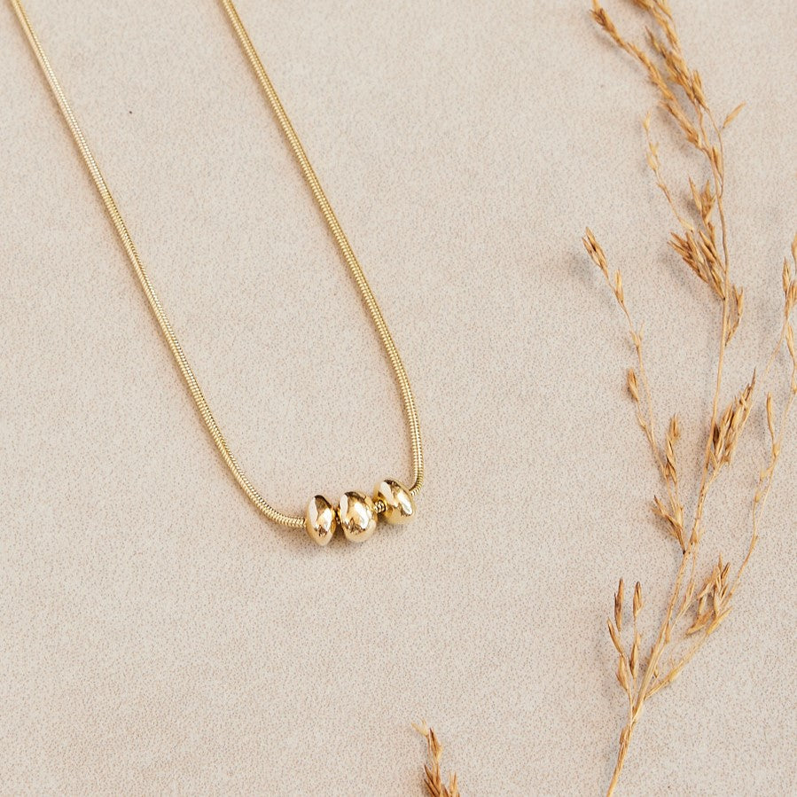 14 karaat gouden hanger Essentials: 3 minimalistische organische gouden steentjes aan een ketting. Liggend op een stenen ondergrond met een gedroogd takje ernaast.