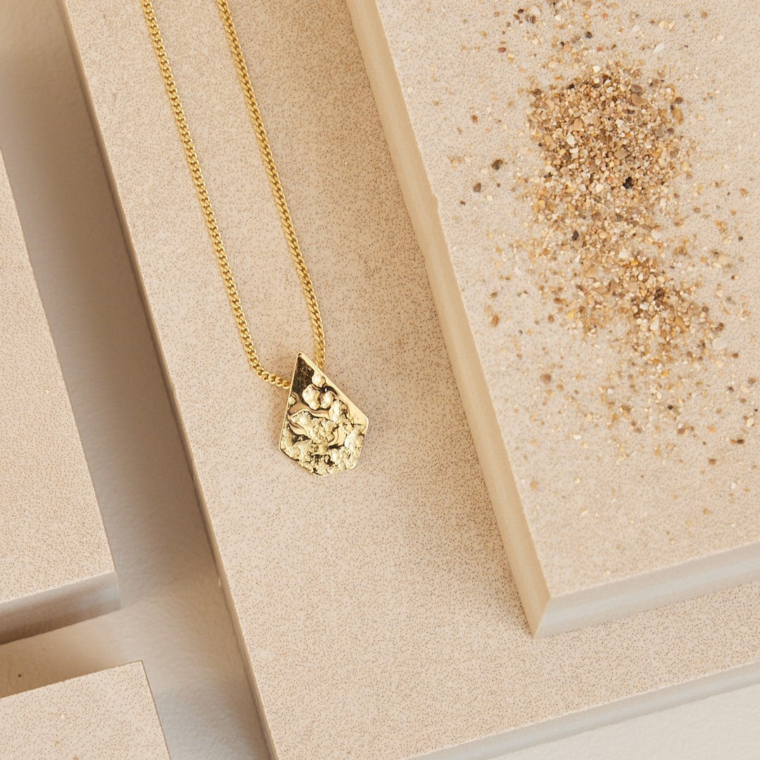 18 karaat geelgoude "Imperfectie Rocks" hanger aan ketting Hanger in de vorm van een steen met zandafdruk erin gewalst voor de ruwe structuur. Liggend op een steen met wat zand eromheen.
