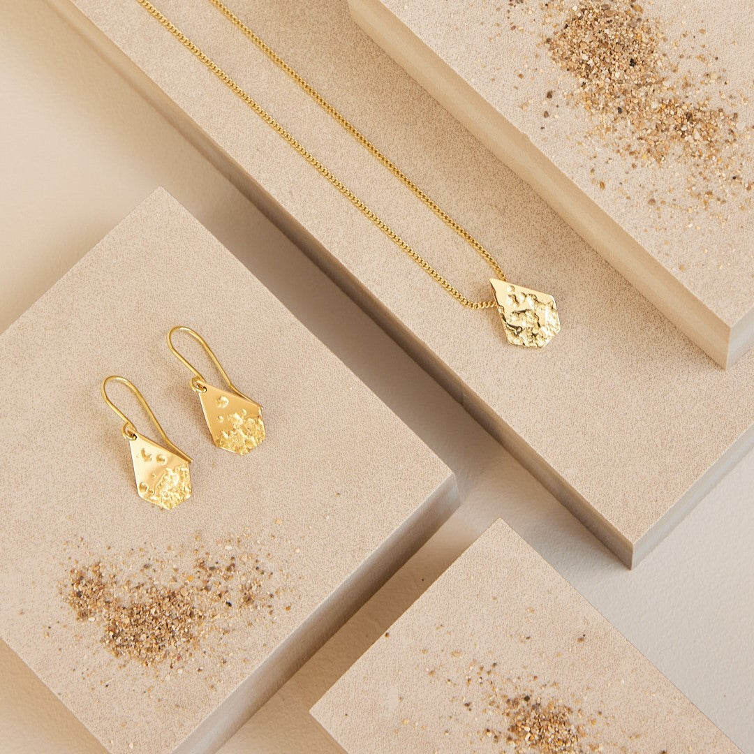 Overzichtsfoto van de collectie Imperfectie Rocks, sieraden met betekenis namelijk dat je goed bent zoals je bent. 18 k gouden hanger en 18 k gouden oorbellen liggend op stenen met wat zand eromheen.