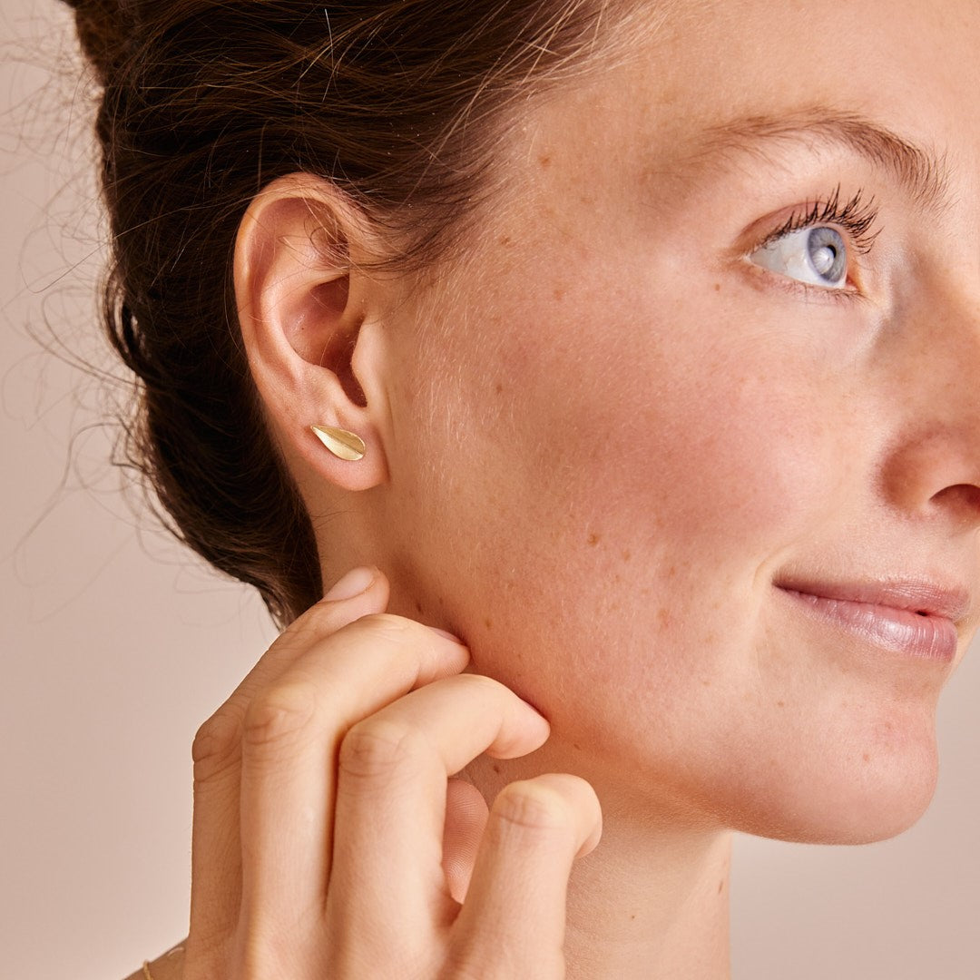 Zijaanzicht van het gezicht van model dat oorsteker Leaves draagt. Een 14 karaat gouden minimalistische blaadje. Hand bij haar kaak.