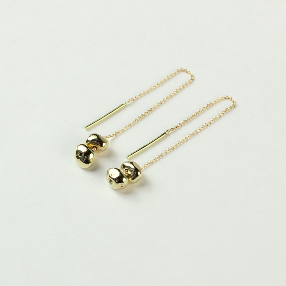 Schuin vooraanzicht van doorrijgoorbellen Oorhangers Essentials: 14 k gouden minimalistische steentjes aan een kettinkje welke je door je oren kan trekken. Liggend op een witte achtergrond.