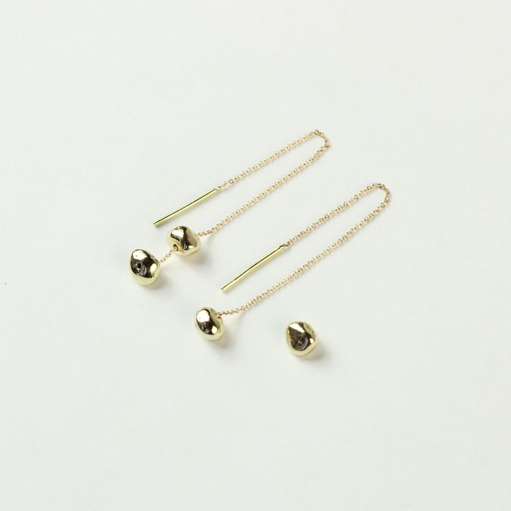 Schuin vooraanzicht van doorrijgoorbellen Oorhangers Essentials: 14 k gouden minimalistische steentjes aan een kettinkje welke je door je oren kan trekken. De bovenste steentjes liggen hier los van het het onderste steentje zodat je kan zien dat je deze van het kettinkje kan halen waardoor ze ook met een enkel steentje te dragen zijn. Liggend op een witte achtergrond.