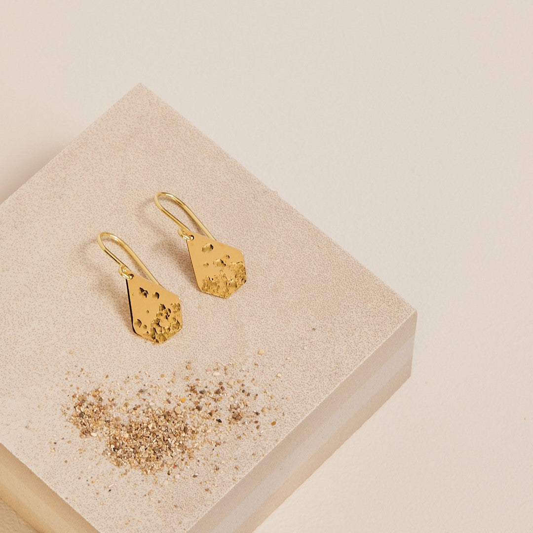 Sfeerfoto van een paar 18 k gouden oorbellen Imperfectie rocks, waar een zandstructuur in gewalst is. De oorhangers liggen op een steen met wat zand eromheen.